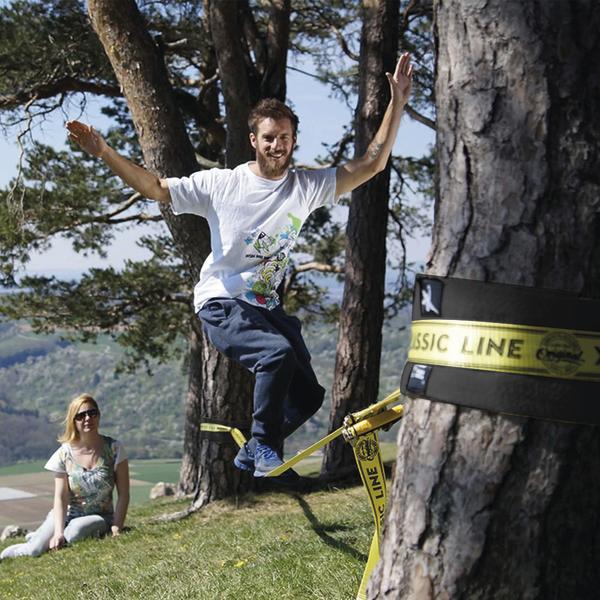 Mann balanciert auf Slackline zwischen Bäumen, Frau sitzt im Gras und schaut zu.