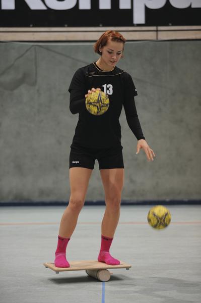 Frau balanciert auf Wackelbrett und hält Handball in Sporthalle.