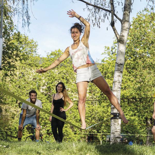 Frau balanciert auf Slackline im Park, zwei Personen beobachten sie im Hintergrund.