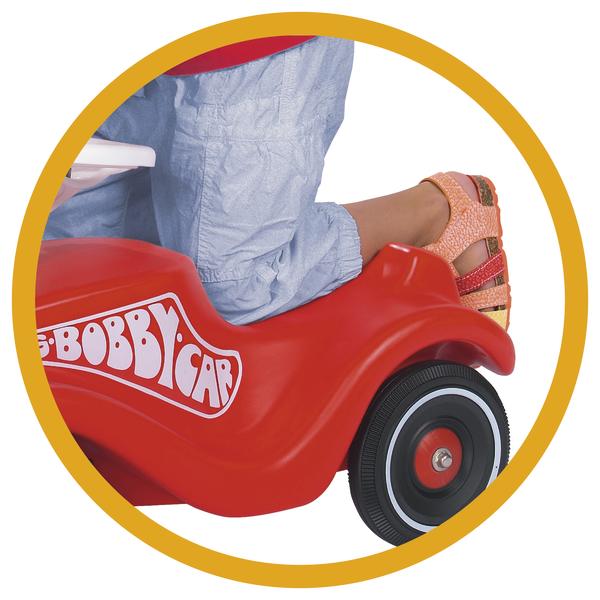 Kind sitzt auf rotem Bobby-Car Rutscher mit schwarzen Reifen und trägt orangefarbene Sandalen.