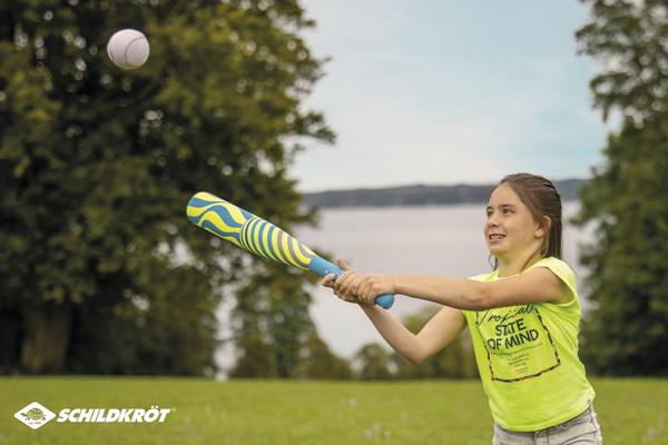 Mädchen schlägt mit buntem Baseballschläger Ball im Park vor See und Bäumen.