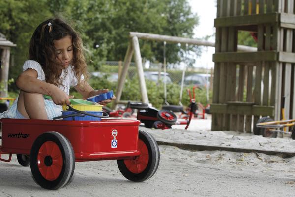 Kind spielt mit Sandspielzeug in rotem Bollerwagen auf Spielplatz im Freien.