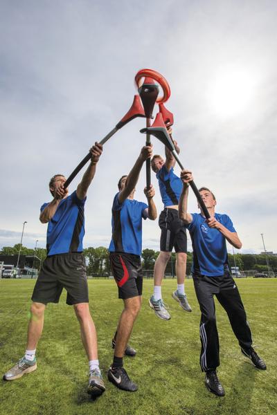 Vier Männer in blauen Trikots spielen auf einem Sportfeld mit großen Lacrosse-Schlägern.