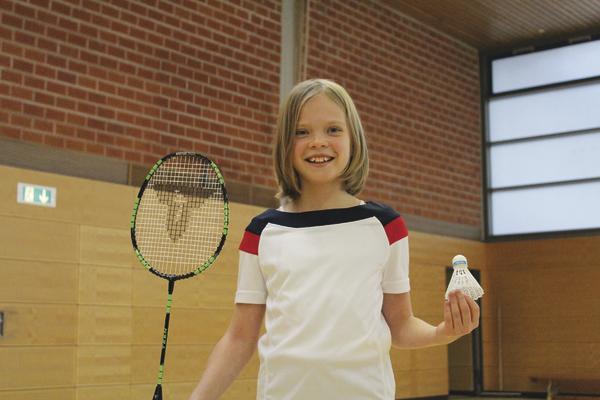 Kind mit Badmintonschläger und Federball in Sporthalle, lächelnd und bereit zum Spiel.