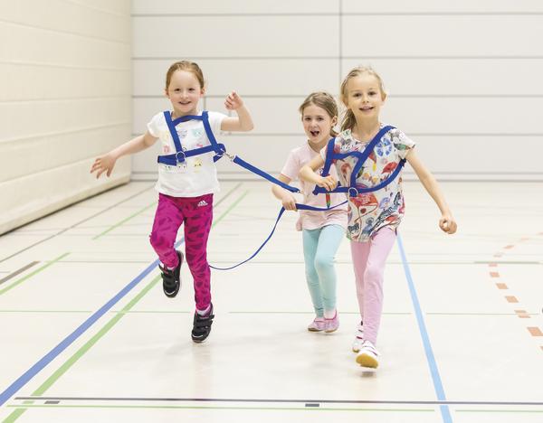 Drei Mädchen laufen mit verbundenen Gurten in einer Sporthalle, spielen kooperatives Bewegungsspiel.