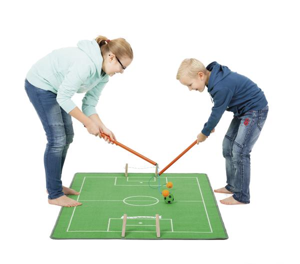 Kinder spielen Tischhockey auf grünem Spielfeld mit orangefarbenen Schlägern und Bällen.