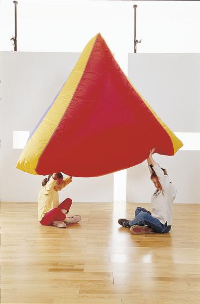 Kinder spielen mit großem, dreieckigem, rotem und gelbem Kissen auf hellem Holzboden.