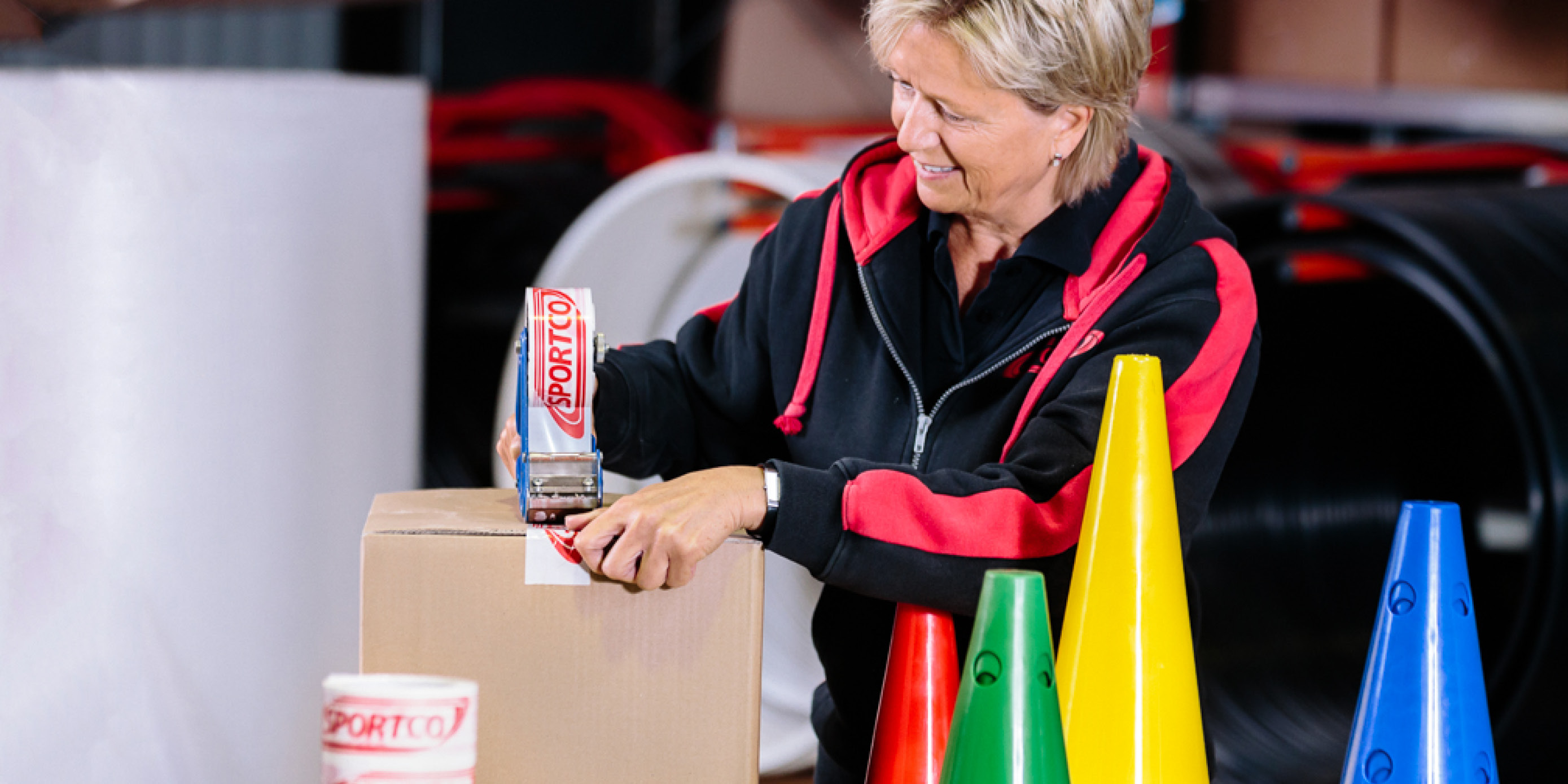 Sportco-Lager-Mitarbeiterin verpackt Ware in einen Karton. Im Vordergrund stehen bunte Markierungskegel.