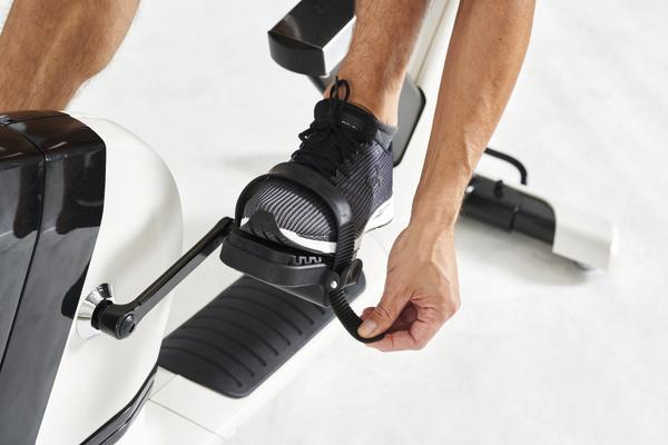 Mann schnallt Fuß in Pedal eines Heimtrainers mit schwarzem Sportschuh und weißer Sohle.