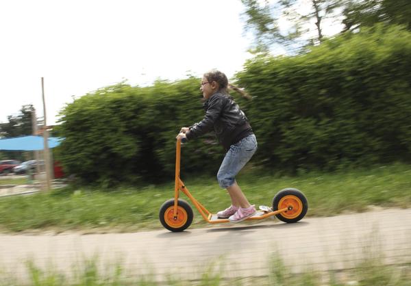 Kind fährt schnellen orangenen Roller auf Gehweg im Park bei sonnigem Wetter.