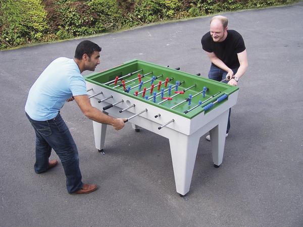 Zwei Männer spielen draußen an einem Tischkicker auf Asphaltfläche, umgeben von Grün.